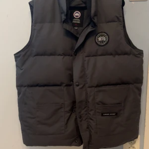 Mörkgrå dunväst från Canada Goose - Snygg mörkgrå dunväst från Canada Goose med hög krage och två stora fickor framtill. Västen har diskret logga på bröstet och stängs med både dragkedja och knappar. Perfekt för lager-på-lager under kyliga dagar. Storleken passar allt från m-xl, vid snabb affär kan pris diskuteras 