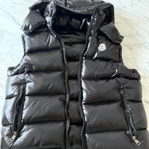 Svart dunväst från Moncler - Svart, glansig dunväst från Moncler med hög krage och två dragkedjefickor framtill. Västen har quiltad design, Moncler-logga på bröstet och innerfoder med serietidningsmönster. Perfekt för lager-på-lager och streetstyle.