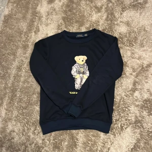 Mörkblå Polo Bear tröja Ralph Lauren - Säljer en mörkblå crewneck från Ralph Lauren med ikoniska Polo Bear-trycket framtill. Tröjan har rund hals, långa ärmar och ribbade muddar. Perfekt för dig som gillar klassisk streetstyle med en twist.