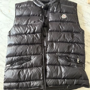 Svart dunväst från Moncler - Snygg svart dunväst från Moncler med quiltad design och hög krage. Västen har två dragkedjefickor framtill och klassisk Moncler-logga på bröstet. Perfekt för lager-på-lager och ger en clean look till din outfit.