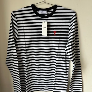 Randig långärmad tröja från Ami Paris  - Säljer en svartvit randig långärmad tröja från Ami Alexandre Mattiussi. Tröjan har rund hals, broderad röd hjärtlogga på bröstet och är gjord i 100% bomull. Perfekt för dig som gillar stilren design med en twist.