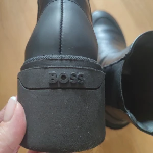 Svarta BOSS boots i skinn - Snygga svarta boots från BOSS med rund tå och platt sula. Tillverkade i slätt skinn med grov klack och diskret logga på hälen. Dragflik baktill i svart och vitt för enkel påtagning. Perfekta för dig som gillar stilrena och robusta skor.