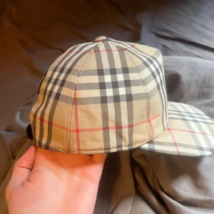 Burberry rutig keps i beige och svart - Snygg Burberry keps med klassiskt rutigt mönster i beige, svart, vitt och röda linjer. Kepsen har böjd skärm och justerbar rem bak. Tillverkad i bomull med mjukt svart innerfoder. Perfekt för dig som gillar ikoniska designeraccessoarer.