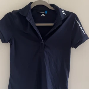 Marinblå piké från J.Lindeberg - Snygg marinblå pikétröja från J.Lindeberg med korta ärmar och klassisk krage. Diskret logga på bröstet, axeln och ryggen. Perfekt för en sportig och stilren look. Tillverkad i mjuk bomull som känns skön mot huden.