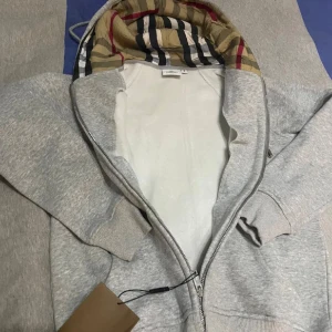 Grå hoodiejacka från Burberry - Snygg grå hoodiejacka från Burberry med dragkedja och klassiskt rutigt foder i huvan. Jackan har ribbade muddar, snörning i huvan och detaljer i silver. Perfekt för dig som vill ha en stilren och lyxig streetwear-look.