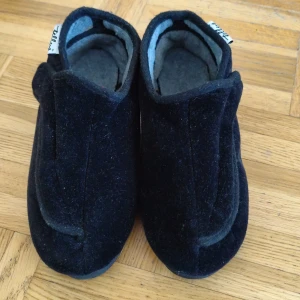 Svart ulltofflor från Tellus - Mysiga svart tofflor från Tellus i mjuk ull med grått foder. De har en enkel och stilren design med praktisk kardborreknäppning för enkel av- och påtagning. Perfekta för att hålla fötterna varma hemma under kyliga dagar.