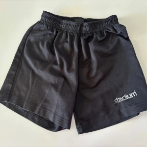 Svarta träningsshorts från Stadium - Svarta träningsshorts från Stadium med elastisk midja och logga tryckt på ena benet. Klassisk och enkel design i mjukt syntetmaterial som passar perfekt för sport eller gym. Lätta och bekväma att bära.
