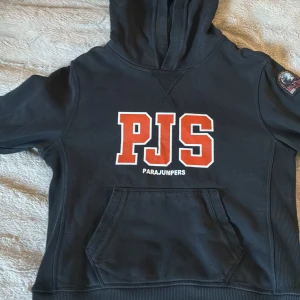 Svart hoodie från Parajumpers - Svart hoodie från Parajumpers med stor röd PJS-logga på bröstet och patch på ärmen. Klassisk känguruficka framtill, huva med dragsko och ribbade muddar. Perfekt för dig som gillar streetwear och vill ha en snygg och bekväm tröja. Perfekt för personer som är mellan 155 till 165 