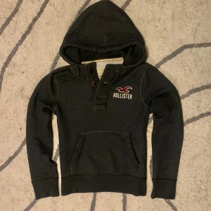 Mörkgrå hoodie från Hollister - Snygg mörkgrå hoodie från Hollister med huva och knappar vid halsen. Tröjan har en klassisk magficka och broderad Hollister-logga i vitt och rött på bröstet. Perfekt för chill dagar och enkel att matcha med jeans eller joggers.