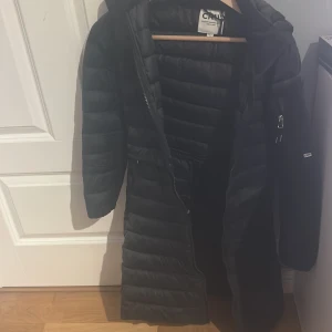 Svart lång dunjacka från CRW - Lång svart dunjacka från Crw med huva och dragkedja framtill. Jackan har quiltad design, tryckknappar i sidorna och en ficka med dragkedja på ärmen. Perfekt för kalla dagar. 