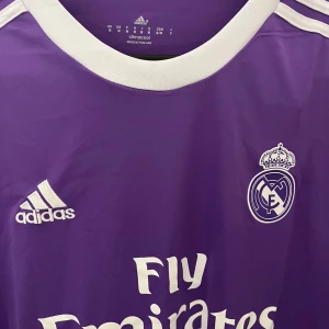 Lila Real Madrid långärmad fotbollströja - Säljer en lila långärmad Real Madrid fotbollströja från Adidas med vitt klubbmärke, Adidas-logga och Fly Emirates-tryck. Tröjan har vita detaljer vid krage och axlar samt är tillverkad i lätt och ventilerande climacool-material.