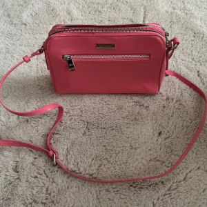 Rosa axelväska från Don Donna - Snygg rosa axelväska från Don Donna med justerbar rem och flera dragkedjefack. Perfekt storlek för mobilen, plånboken och andra småsaker. Väskan har en stilren design och är tillverkad i syntetmaterial.