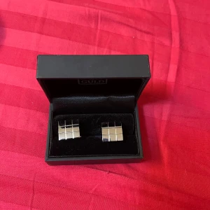 Silvriga manschettknappar Hallbergs Guld - Snygga silvriga manschettknappar från Hallbergs Guld i klassisk fyrkantig form med rutigt mönster. Kommer i original ask, perfekta för att ge din skjorta en stilren och modern touch. Metallfinishen är blank och elegant.