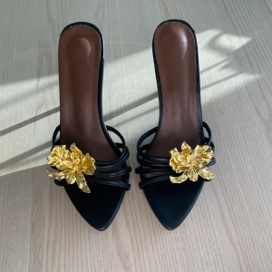 Svarta pumps med guldfärgad blomma - Snygga svarta pumps med spetsig tå och hög klack. Dekorerade med en stor guldfärgad blomma framtill och tunna remmar över foten. Perfekta för dig som vill sticka ut med en unik detalj. Mjuk sula som gör att skon blir väldigt bekväm. Aldrig använda då de är försmå