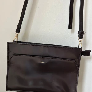 Mörkbrun axelväska från Wera - Snygg mörkbrun axelväska från Wera i slätt skinn med justerbar rem och guldfärgade metalldetaljer. 