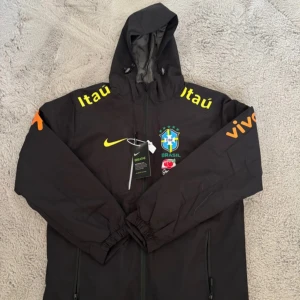 Svart Nike Brasil vindjacka med huva Storlek XS - Svart Nike vindjacka med Brasil-logga, gula och orange tryck på ärmar och bröst, samt huva. Jackan har dragkedja framtill, två fickor och är fodrad med grått tyg. Perfekt för dig som gillar fotboll och vill ha en sportig stil.