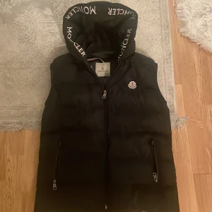 Svart dunväst med huva från Moncler - Svart dunväst från Moncler med broderad logga på bröstet och stor huva med Moncler-tryck. Västen har två dragkedjefickor framtill och hel dragkedja. Insidan har en röd dragkedjeficka och detaljerad etikett. Perfekt för lager-på-lager under kyliga dagar.