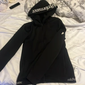Svart vindjacka Peak Performance M - Svart vindjacka från Peak Performance med huva och vit logga på ärmen samt text på huvan. Jackan har dragkedja framtill och fickor på sidorna. Perfekt för dig som gillar stilren och sportig look.