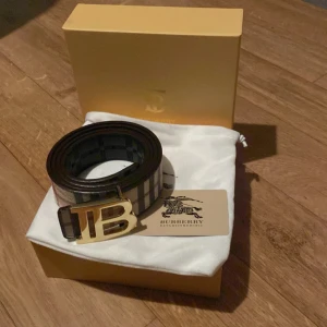 Burberry rutigt skärp med guldspänne - Snyggt Burberry-bälte i klassiskt rutigt mönster med svart och beige bas. Skärpet har ett stort, guldigt TB-spänne framtill och är gjort i äkta skinn. Perfekt accessoar för att lyfta din outfit med en lyxig touch och ikonisk design.priset ej hugget i sten