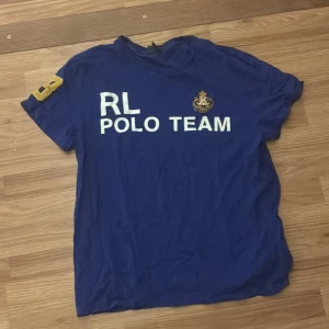 Blå Polo Ralph Lauren t-shirt L - Blå t-shirt från Polo Ralph Lauren i storlek L med vit text 'RL POLO TEAM' på framsidan och ett broderat emblem. Kortärmad modell med gul detalj på ena ärmen. Tillverkad i 100% bomull och har en normal passform. Aldrig använd 