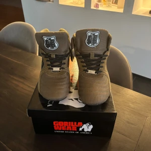 Gorilla Wear Perry High Tops Pro Army Green - Säljer ett par snygga high top sneakers från Gorilla Wear i färgen army green. Använda två eller tre gånger. Skorna har snörning, rund tå och platt sula. Materialmix av mocka och syntet med broderad gorilla på sidan och logga på plösen. Perfekta för gymmet eller streetwear.