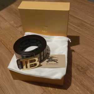 Burberry bälte med guldspänne - Snyggt Burberry bälte i klassiskt rutigt mönster med svart och beige bas, detaljer i brunt och vitt. Bältet har ett stort, guldigt TB-spänne framtill och är gjort i skinn. Perfekt accessoar för att lyfta din outfit med en lyxig touch.