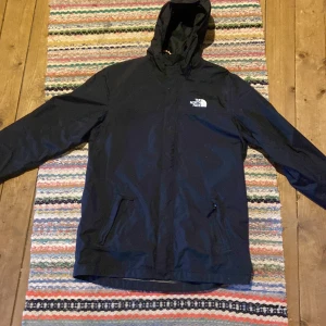 Svart regnjacka The North Face XXL - Svart regnjacka från The North Face i modell Antora, med huva och dragkedja framtill. Jackan har två fickor med dragkedja och klassisk logga på bröstet. Perfekt för blöta dagar och enkel att matcha med allt. Snygg och funktionell design.xxl i barn storlek=small. Den har ett pyttelitet hål i fickan som inte märks av!
