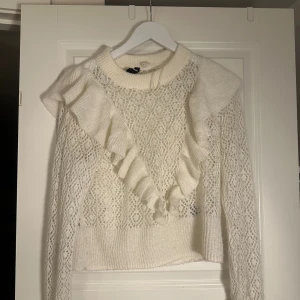 Vit stickad tröja med volang från H&M - Säljer en vit stickad tröja från H&M med snygga volangdetaljer framtill och genombrutet mönster. Tröjan har långärmade ärmar och ribbad kant vid hals och nederdel. Perfekt för dig som gillar romantisk och trendig stil.