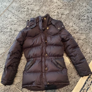 Marinblå dunjacka från Boomerang XS - Säljer en marinblå dunjacka från Boomerang i storlek XS. Jackan har en hög krage, knappar framtill och två snedställda fickor. Perfekt för kalla dagar med sin vadderade och puffiga design. Klassisk och stilren modell som håller dig varm hela vintern.