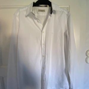 Vit skjorta från Burberry London - Klassisk vit skjorta från Burberry London med diskret rutigt tyg på insidan av kragen. Skjortan har lång ärm, knappar framtill och stilren passform. 