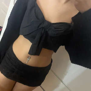 Svart croppad tubtopp med rosett - Supersnygg svart croppad tubtopp med stor rosett framtill och knyt-detalj. Axelbandslös design som sitter tight och framhäver midjan. Perfekt för dig som vill sticka ut med en cool och trendig look. Materialet är lätt och känns skönt mot huden.