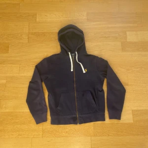 Mörkblå hoodie från Polo Ralph Lauren - Mörkblå hoodie från Polo Ralph Lauren med dragkedja, vit snörning i huvan och klassisk gul broderad logga på bröstet. Tröjan har känguruficka framtill och är tillverkad i mjuk bomull. Perfekt för chill dagar och snygg streetstil.