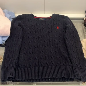 Mörkblå kabelstickad tröja Polo Ralph Lauren - Kabelstickad mörkblå tröja från Polo Ralph Lauren med rund halsringning och röd broderad logga på bröstet. Tröjan har långa ärmar och ribbade muddar för en klassisk look. Perfekt för lager-på-lager under kyliga dagar.