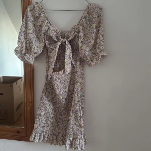 Blommig kortklänning med puffärm - Supersöt kortklänning med blommigt mönster i pastelliga färger. Klänningen har puffiga kortärmar, volangkant nertill och en snygg knytning framtill. Materialet känns lätt och luftigt, perfekt för varma dagar. Öppen detalj vid magen för extra sommarkänsla.