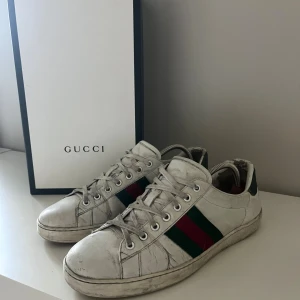 Gucci Ace vita sneakers - Gucci Ace sneakers i vitt skinn med klassiska gröna och röda ränder på sidorna och gröna detaljer på hälen. Låga sneakers med snörning och rund tå. Kommer med originalkartong, dustbag och äkthetsbevis. Perfekta för dig som vill ha en ikonisk och stilren look.  (Hör gärna av dig vid minsta funderingar)☺️