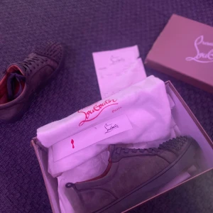 Christian Louboutin svarta sneakers med nitar - Säljer ett par svarta sneakers från Christian Louboutin i mocka med coola svarta nitar på tån. Skorna har snörning och en klassisk låg siluett. Insidan är röd, vilket ger en lyxig känsla. Kommer med originalkartong och dustbag.