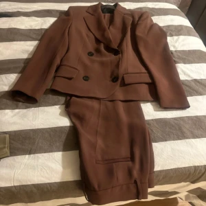 Brun kostym från Zara, storlek S - Säljer en snygg brun kostym från Zara i storlek S. Kavajen är dubbelknäppt med svarta knappar och har klassiska slag samt fickor med lock. Byxorna har raka ben och pressveck. Perfekt för dig som vill ha en stilren och trendig look.
