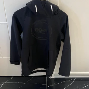 Svart hoodie från 8848 Altitude - Svart hoodie från 8848 Altitude med stor huva och vita snören. Tröjan har ett diskret tryck på bröstet, långa ärmar och praktiska fickor med dragkedja. Perfekt för dig som gillar en clean och sportig look.