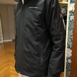 Svart vindjacka Peak Performance - Svart vindjacka från Peak Performance med huva och dragkedja framtill. Jackan har flera fickor med dragkedja, diskret logga på bröstet och ärmarna samt en clean, sportig look. Perfekt för dig som gillar stilren och funktionell design. Den funkar som vinter jacka för den är fodrad med. Storlek S