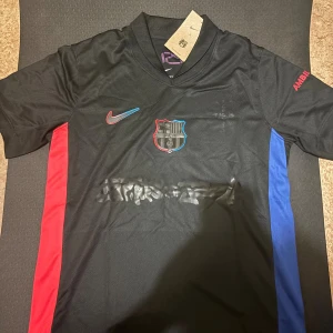 Barcelona x Travis scott fotbollströja - Snygg svart FC Barcelona fotbollströja från Nike med klubbmärke på bröstet, röd och blå detalj längs sidorna och broderad Nike-logga. Tröjan har korta ärmar och är tillverkad i lätt, ventilerande material som passar perfekt för match eller träning.
