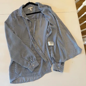 Randig skjorta från H&M - Snygg skjorta från H&M med blå och vita vertikala ränder. Klassisk krage, knäppning framtill och långa ärmar med manschett. Skjortan har fint fall och har en relaxed fit men ändå blir välklätt.