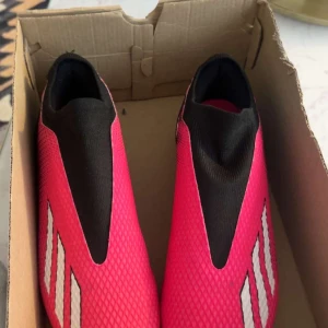 Adidas rosa och svarta fotbollsskor - Säljer ett par Adidas fotbollsskor i en riktigt snygg rosa och svart färgkombination. Skorna har en strumpliknande passform utan snörning, med vit Adidas-logga på sidan och mönstrad ovandel i syntetmaterial. Sulan är rosa med fasta dobbar för gräsplan.