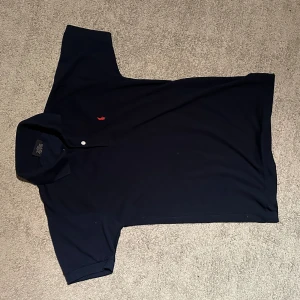 Mörkblå pikétröja från Ralph Lauren - Klassisk mörkblå pikétröja från Ralph Lauren med röd broderad logga på bröstet. Tröjan har korta ärmar, krage och två knappar framtill. Tillverkad i mjuk bomull som känns skön mot huden. Perfekt för en clean och stilren look.