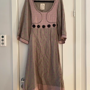 Blommig långklänning från Noa Noa - Långklänning i beige med rosa blommönster från Noa Noa. Klänningen har vida trekvartsärmar, dekorativa svarta pärlblommor vid bröstet och ljusrosa detaljer med grönt mönster vid halsringning, ärmslut och nederkant. Tillverkad i mjuk viskos, perfekt för dig som gillar boho-vibe. Mått Över bröstet: ca 52 cm Längd från axeln och ner: ca 112 cm  100% viskos  Kommer från ett djur och rökfritt hem. 