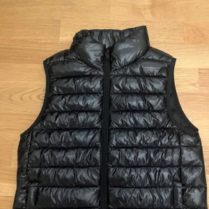 Svart dunväst med dragkedja - Säljer en svart dunväst med hög krage och horisontella sömmar. Västen har en stilren look med hel dragkedja framtill och är lätt vadderad för extra värme. Perfekt att slänga över en hoodie eller tröja när det är kyligt ute.