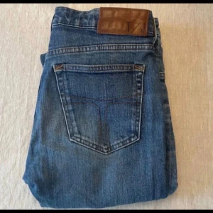 Blå jeansbyxor från T/J 27/32  - Snygga blå jeansbyxor från T/J med klassisk femficksdesign och brunt läderpatch bak i midjan. Jeansen har raka ben och är tillverkade i slitstarkt denimtyg med subtila tvättade detaljer. Perfekt för en avslappnad och cool stil.