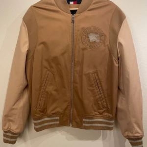 Tommy Hilfiger brun collegejacka bomberjacka - Brun Tommy Hilfiger bomberjacka med en broderad logotyp på bröstet. Jackan har en dragkedja fram, ribbade muddar och en klassisk bomberjacka-silhuett. Perfekt för en avslappnad men stilfull look.