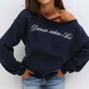 Mörkblå offshoulder hoodie med text - Snygg mörkblå hoodie med offshoulder-design och vit text 'Dansar vidare i livet' på bröstet. Tröjan har en stor magficka och långa ärmar med ribbade muddar. Perfekt för dig som gillar en avslappnad och trendig stil.