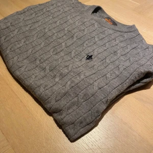 Grå kabelstickad tröja från Morris - Säljer en grå kabelstickad tröja från Morris med rund halsringning och diskret broderad logga på bröstet. Tröjan har lång ärm och är tillverkad i mjukt material som passar perfekt för kyligare dagar.