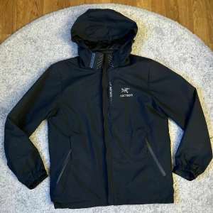 Svart vindjacka från Arc'teryx - Svart vindjacka från Arc'teryx med avtagbar huva, dragkedja och diskreta reflexdetaljer. Jackan har två snedställda fickor med dragkedja och minimalistisk design. Perfekt för dig som gillar stilren och funktionell outdoor-look.